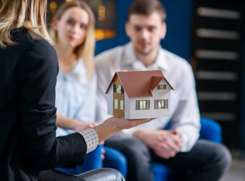 Close up shot of female realtor, interior designer hands showing 3d model of house to cute caucasian couple on the blue modern office background.